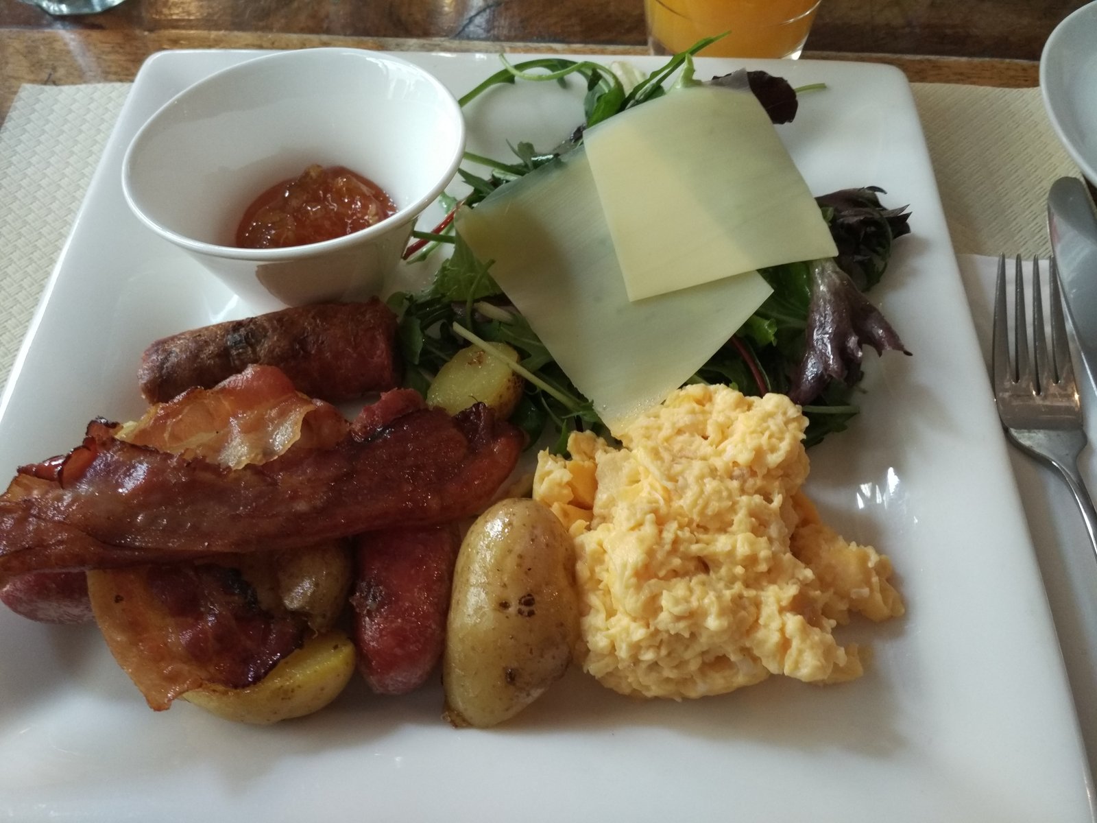 A brunch plate with scrambled eggs, bacon, sausage, potatoes, cheese and greens