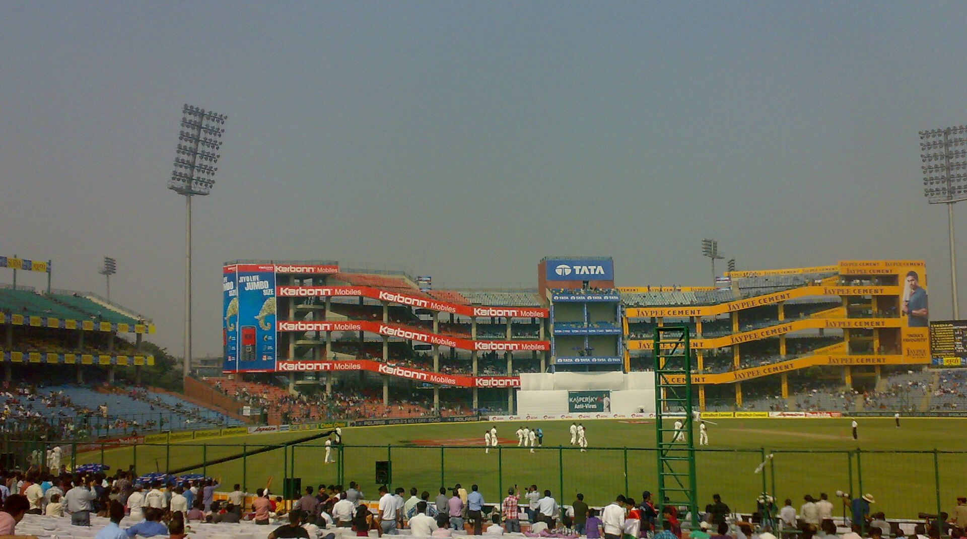 Daytime view of the Feroz Shah Kotla-era stands with Karbonn Mobiles, Tata and Jaypee Cement advertisements, players in white on the field, and tall floodlight pylons.
