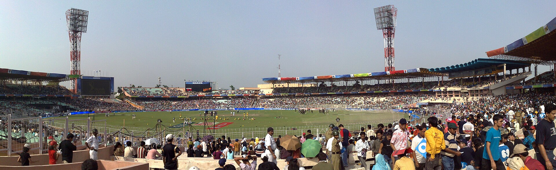 Wide panorama of Eden Gardens on a match day, with a sea of spectators in the foreground stands, the green outfield, and the twin red-and-white floodlight towers at either end.