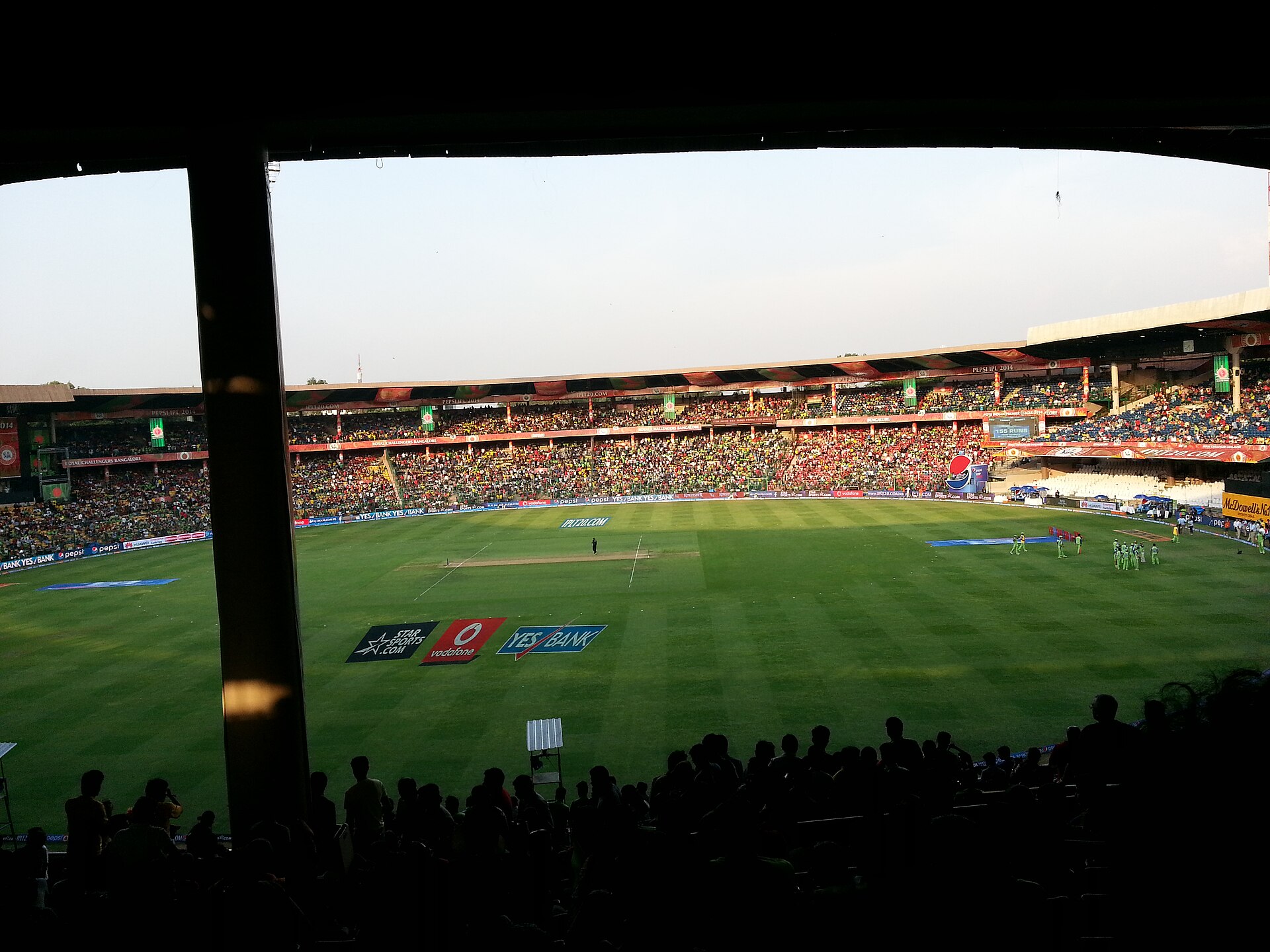 View of the outfield and packed stands during an IPL 2014 match at Chinnaswamy Stadium, framed by a dark stand pillar and roof, with Vodafone, Yes Bank and Star Sports logos painted on the grass.