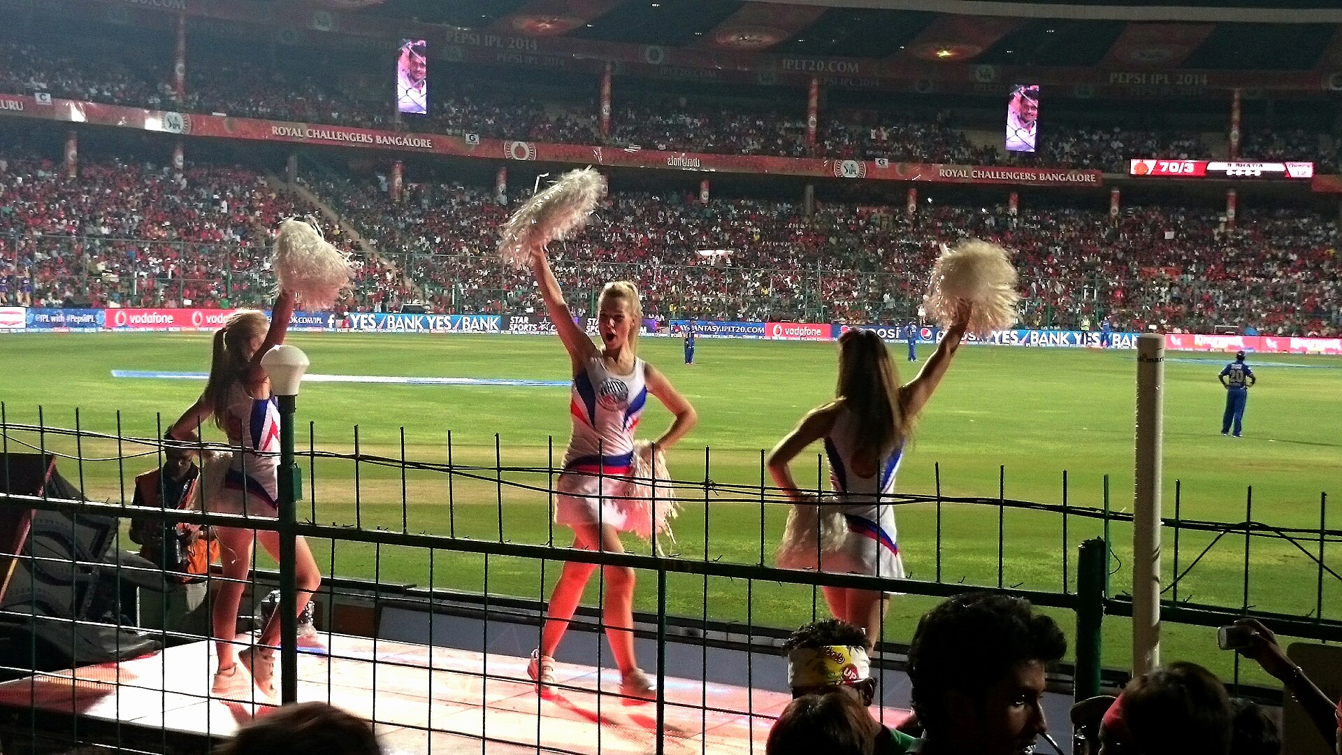 Cheerleaders performing on a boundary-side platform during an IPL night game at Chinnaswamy Stadium, with the packed stands and Royal Challengers Bangalore branding behind them.
