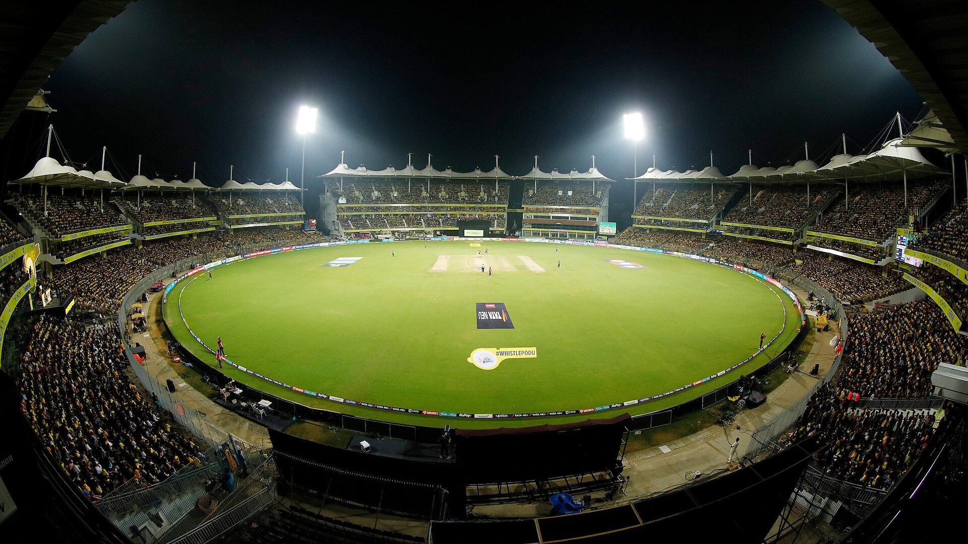 Night view of a packed Chepauk stadium under floodlights during a CSK game, with the glowing green outfield and #WHISTLEPODU and TATA NEU logos painted on the grass.