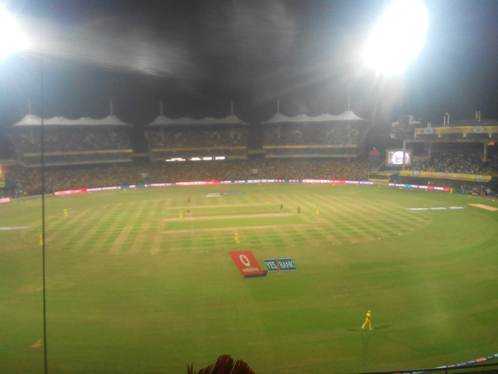 Grainy night photo of an IPL match at Chepauk in 2013, with players in yellow on the outfield, packed stands, floodlights blazing, and Vodafone and Yes Bank logos painted on the grass.