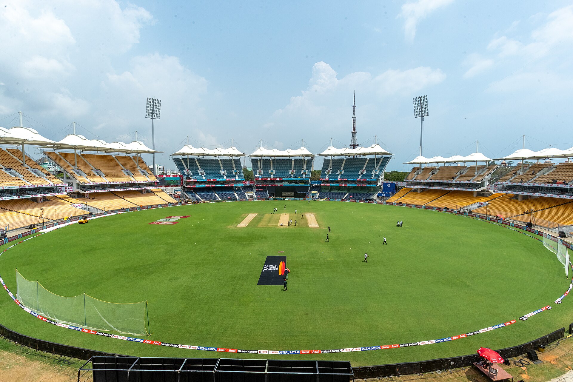Daytime view of M. A. Chidambaram Stadium's oval outfield, with blue and yellow stands under white canopy roofs and two floodlight towers against a cloudy sky.