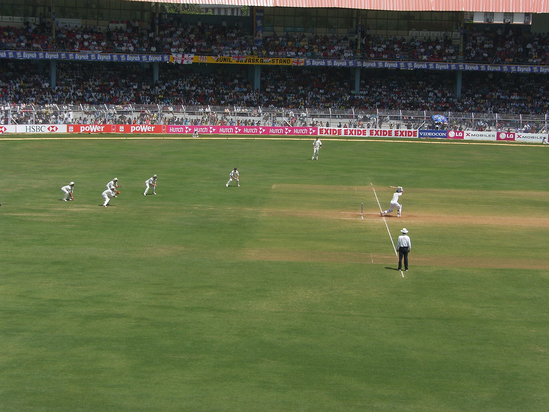 Test match action at Wankhede, with a batsman playing a stroke as fielders in white crouch in the slips, a packed Sunil Gavaskar Stand behind, and HSBC, Hutch and Exide hoardings on the boundary.